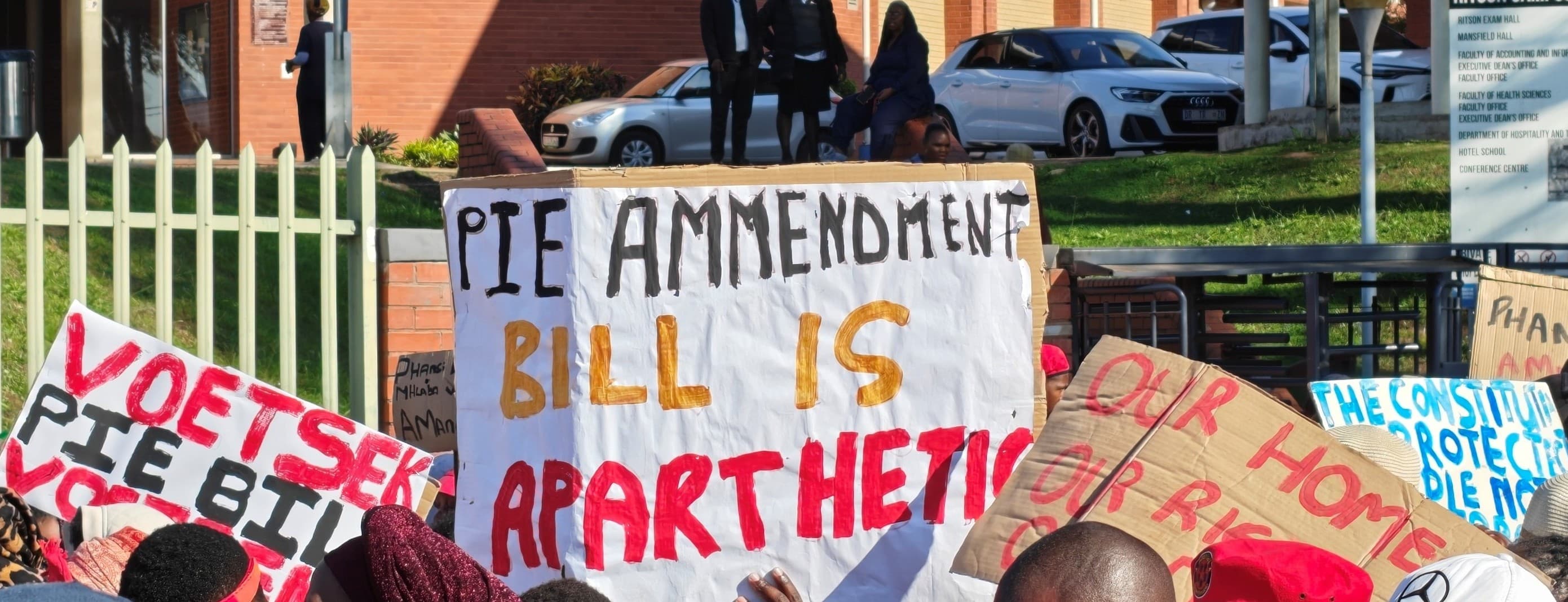 Protesters hold handmade placards outside a red-brick building. The largest reads 'PIE Amendment Bill is apartheid', and others read 'Voetsek PIE Bill' and 'The Constitution protects'.