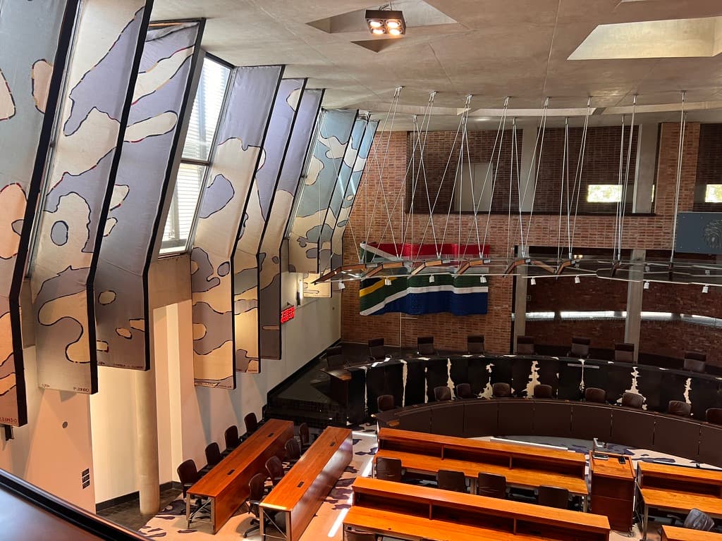 Interior of the Constitutional Court chamber in Johannesburg, seen from the public gallery. Exposed red-brick walls, a large South African flag and angled banners painted with abstract shapes surround a curved judges' bench and rows of wooden tables.