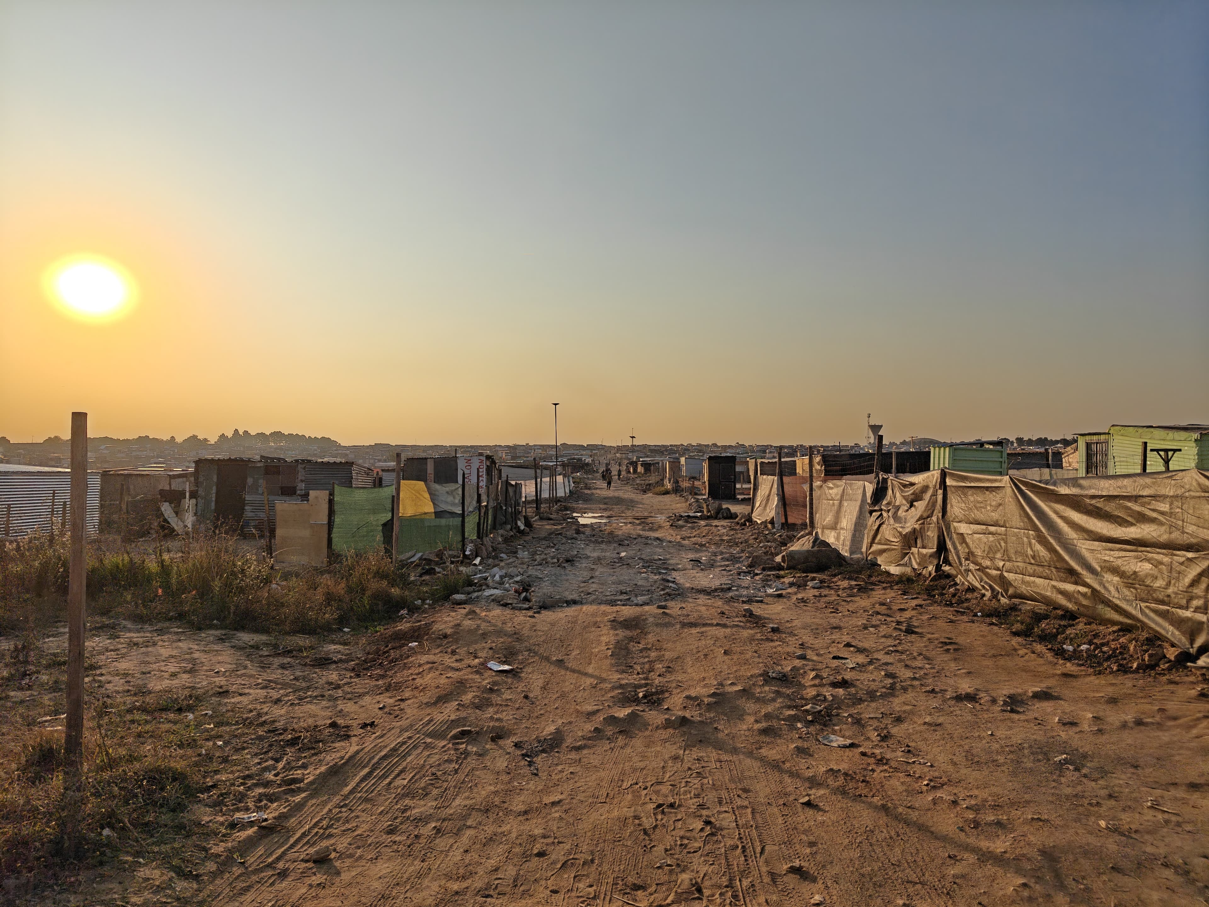 A dirt road runs through the eNkanini informal settlement at sunset, lined on both sides by shacks built from corrugated iron, wooden boards and tarpaulin, with the low sun glowing in a hazy orange sky.